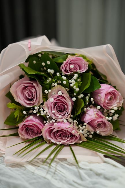 A bouquet of fresh, pastel pink roses with delicate, layered petals arranged with bright green foliage and small white baby's breath flowers. The flowers are wrapped in elegant white tissue paper and